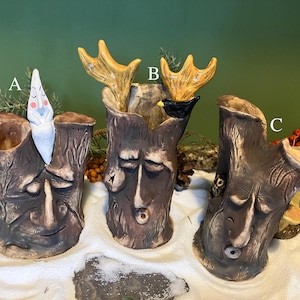 Puede incluir: Tres jarrones de cerámica con forma de tronco de árbol con caras caprichosas. El jarrón de la izquierda tiene una figura de fantasma blanco en la parte superior, el jarrón del medio tiene un par de cuernos y el jarrón de la derecha tiene una pequeña figura de pájaro.
