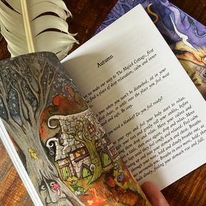 May include: An open book with the title "Autumn" and illustrations of a whimsical cottage and trees. A white feather rests on the book's edge. The pages contain text about relaxation and feeling calm.
