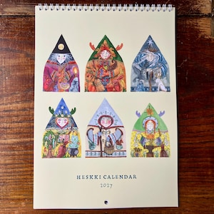 Könnte beinhalten: Ein spiralgebundener Wandkalender mit dem Titel HESKKI CALENDAR 2027 auf cremefarbenem Hintergrund. Das Deckblatt zeigt sechs dreieckige Illustrationen von Figuren in farbenfroher, detaillierter Kleidung, die jeweils in einer Spitzbogenform gerahmt sind und verschiedene saisonale Themen sowie Naturelemente in einem lebendigen, künstlerischen Stil darstellen.