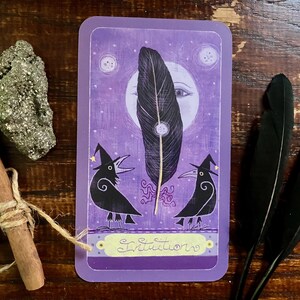 May include: A purple tarot card with a large black feather, two crow illustrations wearing witch hats, and the word "Intuition". A pyrite stone, a cinnamon stick, and two black feathers are also present.