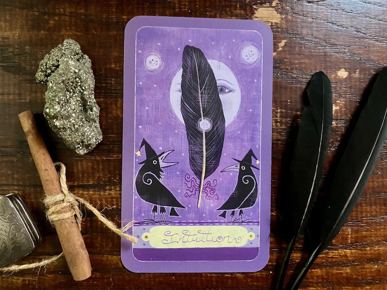 May include: A purple tarot card with a large black feather, two crow illustrations wearing witch hats, and the word "Intuition". A pyrite stone, a cinnamon stick, and two black feathers are also present.
