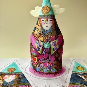 May include: A fabric doll with a teal hat, white wings, and a pink and purple patterned dress. The doll has a painted face and is holding a small animal. The base of the doll has the word "Blessings" on it.