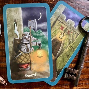 May include: Two tarot cards with blue borders and a decorative axe and key on a wooden surface. One card depicts a guard holding a lantern and axe, with the word "Guard" at the bottom. The other card shows a castle entrance.
