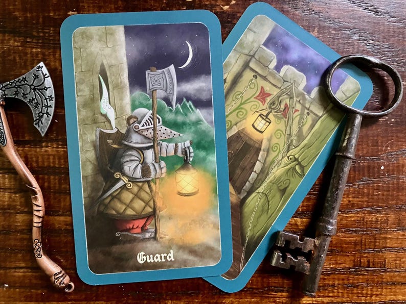 May include: Two tarot cards with blue borders and a decorative axe and key on a wooden surface. One card depicts a guard holding a lantern and axe, with the word "Guard" at the bottom. The other card shows a castle entrance.