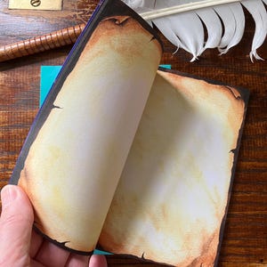 May include: A notebook with pages designed to look like aged parchment. The edges of the pages are darkened, giving the appearance of an antique document. A wooden pen and a feather are also visible.