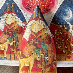 May include: A cone-shaped fabric ornament featuring a printed illustration of a figure with white hair and horns wearing a red coat, holding a wooden staff, and standing with a fawn. The background fabric displays the text BRIGHTEST 2027 BLESSINGS.