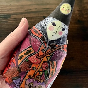 May include: A soft fabric doll shaped like a cone, featuring a printed illustration of a witch with white hair, a black pointed hat with a moon, and a colourful robe. The design includes a broomstick, a purple jack-o'-lantern, and orange accents.
