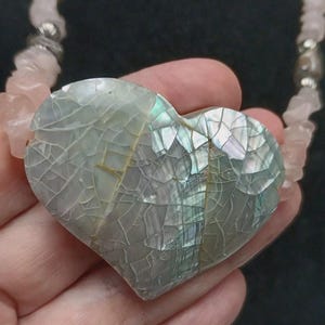 Puede incluir: Un colgante en forma de corazón con un mosaico de fragmentos de concha iridiscentes en tonos grises y verdes, suspendido de un collar compuesto por esquirlas irregulares de cuarzo rosa pálido y pequeñas cuentas espaciadoras plateadas.