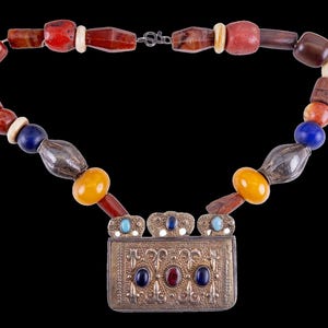 May include: A handcrafted necklace featuring a variety of beads in shades of red, brown, yellow, blue, and white. The necklace includes a silver rectangular pendant with ornate detailing and colorful stone accents. The necklace is displayed against a black background.
