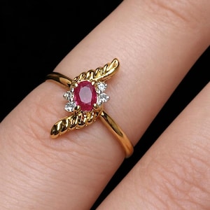 May include: A gold ring featuring a central oval-cut red ruby gemstone flanked by small clear accent stones. The setting includes two textured, wing-like gold elements extending from the sides of the central stone cluster on a thin gold band.