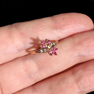 Op de afbeelding: Een goudkleurige ring met een bloemmotief. De bloem is versierd met roze en heldere edelstenen. De ring wordt in een hand gehouden tegen een donkere achtergrond. De ring heeft een dunne en delicate band.