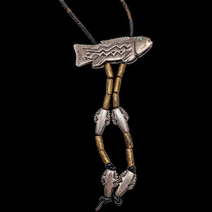 May include: A necklace featuring a silver fish pendant with a small green stone eye, suspended from a black cord. The design includes dangling brass-colored cylindrical beads and four smaller silver fish-shaped charms at the base.