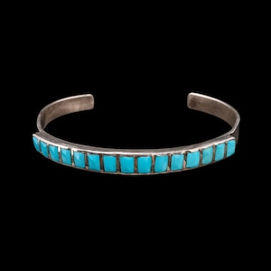 May include: A silver cuff bracelet featuring a row of rectangular, bright blue turquoise stones inlaid along the front. The metal band has a smooth, polished finish and an open back design, allowing for easy adjustment on the wrist.