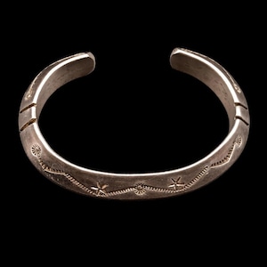 May include: A silver-toned metal cuff bracelet featuring a curved, open-ended band. The surface is decorated with a repeating engraved pattern consisting of small star-like shapes and circular sun motifs connected by a thin, wavy line.