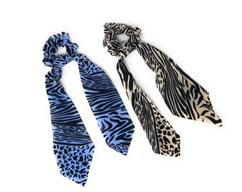 May include: Two hair scrunchies with long tails. One scrunchie is blue with a black and white zebra print and the other is beige with a black and white zebra print.