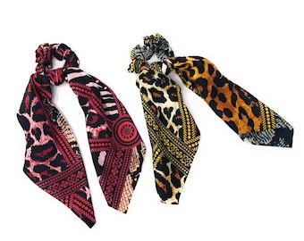 May include: Two colorful hair scrunchies with leopard print and geometric patterns. The scrunchies are made of fabric and have a black background.