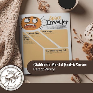 Puede incluir: Un cuaderno de trabajo sobre salud mental infantil titulado "Space Invader" con un personaje de sol de dibujos animados. El cuaderno forma parte de la "Serie de salud mental infantil: Parte 2: Preocupación". La imagen incluye una manta de punto beige, una taza de café y una esfera decorativa.