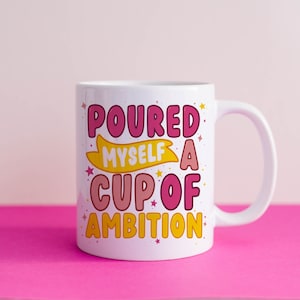 Puede incluir: Taza de cerámica blanca con un diseño rosa, amarillo y blanco. La taza dice "Poured Myself a Cup of Ambition".