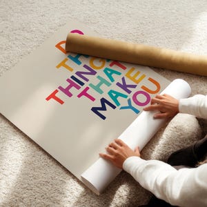 May include: A person unrolling a poster with the text "Do the things that make you happy" in colorful letters. The poster is on a white background.