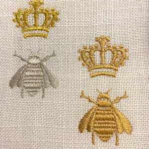May include: A white linen fabric with embroidered gold and silver crowns and bees. The crowns have a pointed top and are decorated with a series of small circles. The bees have a striped body and six legs.