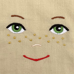 May include: Embroidered face with green eyes, brown eyebrows, a small pink nose, and a red smile. The face has gold freckles on a beige background.