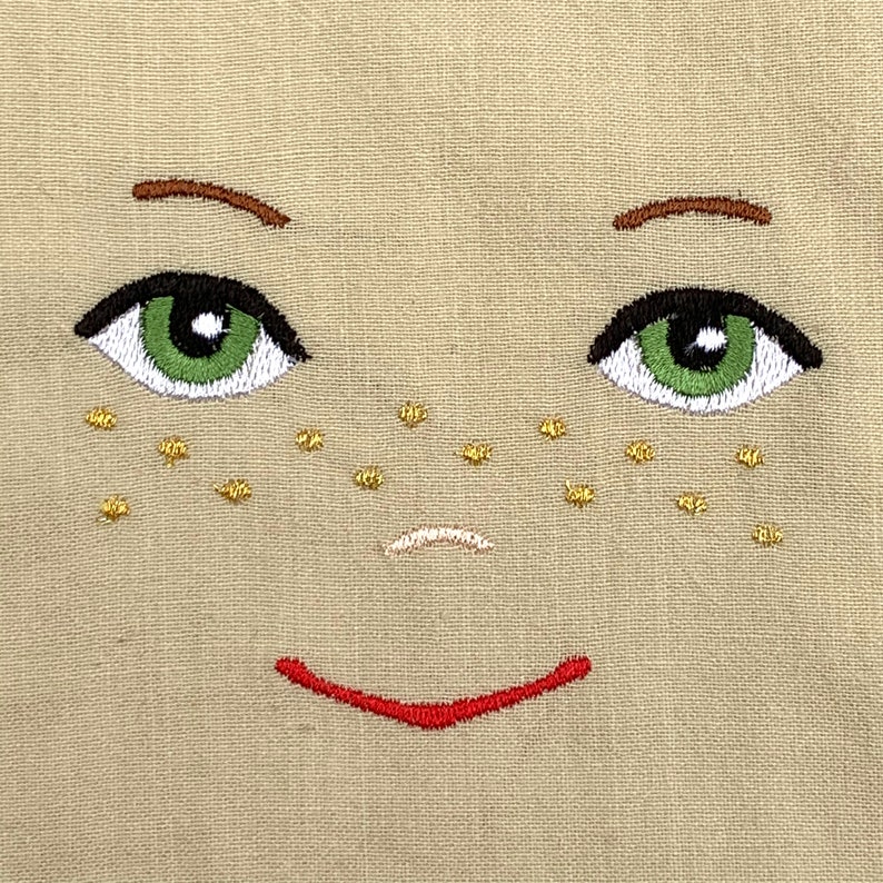 May include: Embroidered face with green eyes, brown eyebrows, a small pink nose, and a red smile. The face has gold freckles on a beige background.