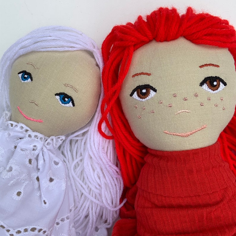 May include: Two handmade fabric dolls, one with white yarn hair and a white dress, and the other with red yarn hair and a red sweater.