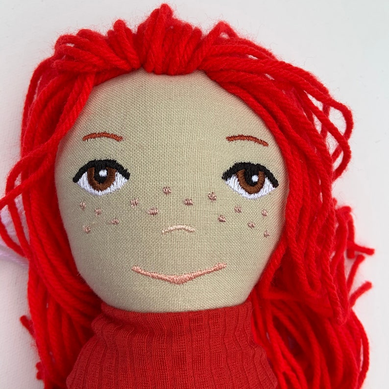 May include: Handmade doll with red yarn hair, embroidered facial features, and a red sweater.