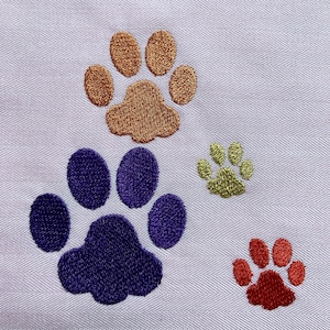 May include: Four embroidered paw prints in different colors: brown, purple, green, and red. The paw prints are on a white fabric background.