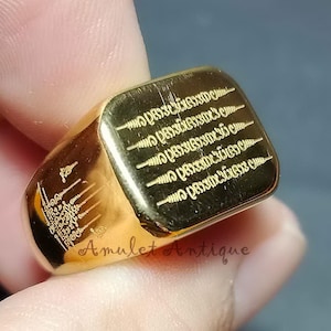 Può includere: Un anello con sigillo color oro con testo e simboli incisi. L'anello ha una faccia rettangolare con iscrizioni dettagliate. Le parole "Amulet Antique" sono visibili sulla superficie dell'anello. L'anello è tenuto da una persona.