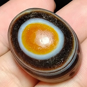 May include: A smooth, oval-shaped agate stone with a distinctive eye pattern. The eye is a concentric circle of white, orange, and brown bands. The stone is dark brown with a glossy finish.