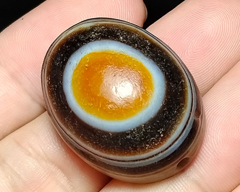May include: A smooth, oval-shaped agate stone with a distinctive eye pattern. The eye is a concentric circle of white, orange, and brown bands. The stone is dark brown with a glossy finish.