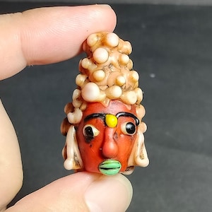 May include: A small, hand-crafted bead shaped like a face with a red base, black eyebrows and eyes, a yellow forehead dot, and green lips. The head is topped with a textured, cream-colored cluster of small, rounded, bubble-like protrusions.