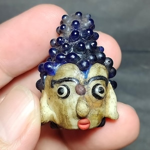 May include: A small, handcrafted glass bead shaped like a stylised head with a textured, dark blue, bumpy crown. The face is off-white with black-outlined eyes, a small brown dot between the brows, and bright red lips, creating a distinct character.