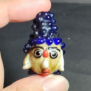 Puede incluir: Una pequeña cuenta de vidrio hecha a mano con forma de rostro estilizado y un sombrero cónico alto, texturizado y de color azul oscuro. El rostro presenta una tez bronceada clara, ojos delineados en negro, una nariz roja y labios rojos.
