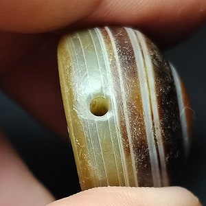 May include: A close-up of a striped agate bead with a hole drilled through the center. The bead is a mix of brown, white, and gray stripes.
