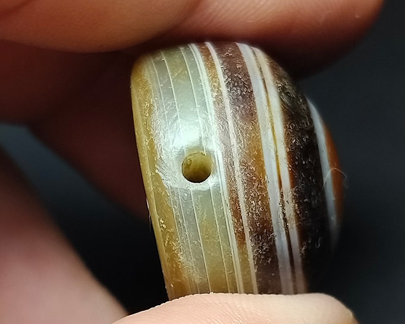 May include: A close-up of a striped agate bead with a hole drilled through the center. The bead is a mix of brown, white, and gray stripes.