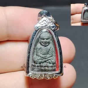 Könnte beinhalten: Ein silberner Anhänger mit einer sitzenden Buddha-Figur in einem klaren, gewölbten Rahmen. Der Buddha ist in einer meditativen Pose dargestellt, mit aufwendigen Details. Der Anhänger hat eine dekorative Öse und einen roten Hintergrund.
