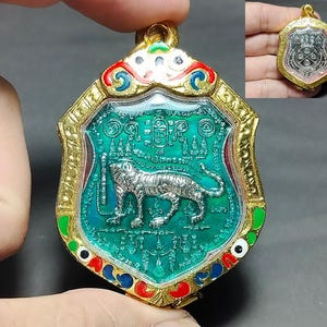 Może przedstawiać: Złoty wisiorek w kształcie tarczy, z srebrnym tygrysem na turkusowym tle. Wisiorek ma skomplikowane detale i kolorowe emaliowane akcenty w kolorze czerwonym, niebieskim i zielonym. Mniejsza wersja pokazana jest w prawym górnym rogu.