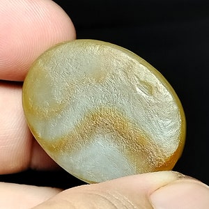 May include: A smooth, oval-shaped, light brown agate gemstone with white and gold swirling patterns.