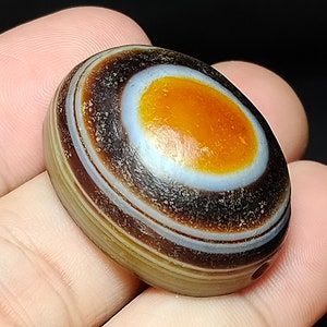 May include: A brown, white, and orange agate stone with a hole drilled through it. The stone has a circular pattern of concentric rings.