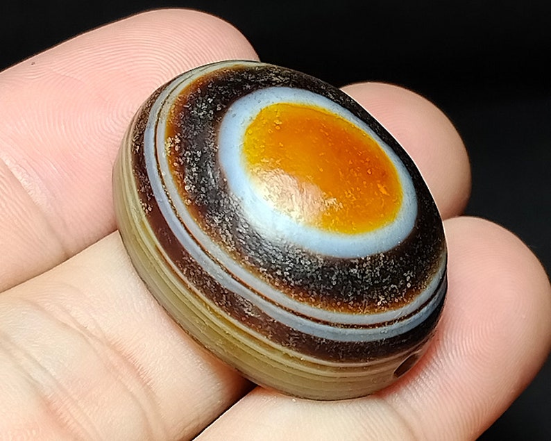 May include: A brown, white, and orange agate stone with a hole drilled through it. The stone has a circular pattern of concentric rings.