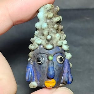 Puede incluir: Una pequeña cuenta de vidrio hecha a mano con forma de rostro estilizado, tez azul oscuro, ojos grandes blancos y negros, boca amarilla brillante y un peinado o tocado cónico texturizado, abultado y de color verde claro.