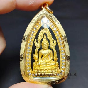 Pode incluir: Pingente dourado com uma figura de Buda sentado dentro de uma moldura transparente em forma de gota. A moldura é adornada com pequenas pedras preciosas transparentes e detalhes dourados intrincados. A estátua de Buda está em pose meditativa.