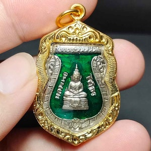 Könnte beinhalten: Ein goldener Anhänger mit einem grünen und silbernen Buddha-Bild im Inneren. Der Anhänger ist verziert und hat ein detailliertes Design. Der Buddha sitzt in meditativer Haltung mit den Händen im Schoß.