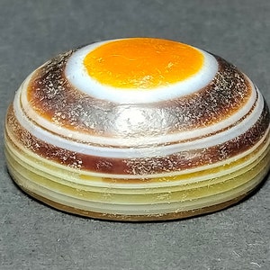 May include: A round, banded agate stone with a white center and an orange circle in the center. The stone has brown, white, and yellow bands.
