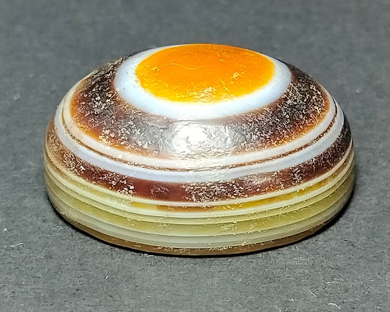 May include: A round, banded agate stone with a white center and an orange circle in the center. The stone has brown, white, and yellow bands.