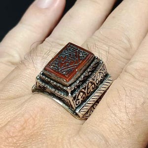 以下が含まれることがあります： 装飾的なデザインと長方形の赤茶色の石が付いたシルバーリング。石には複雑な彫刻が施されています。リングは指にあります。
