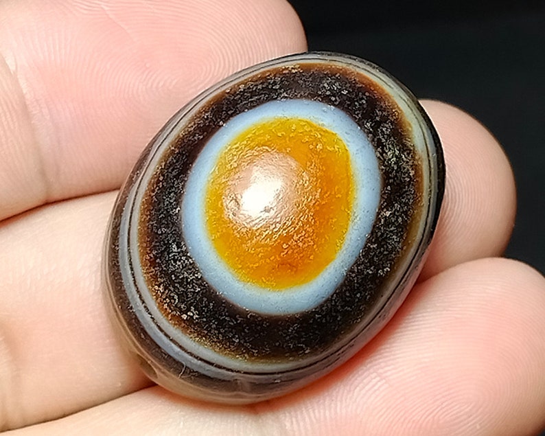 May include: A smooth, oval-shaped agate stone with a distinctive eye pattern. The eye is a concentric circle of white, orange, and brown bands.