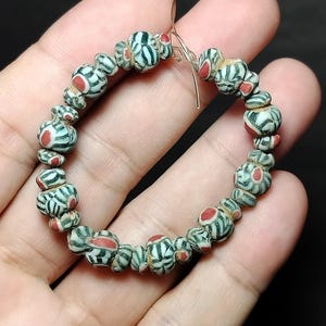 Könnte beinhalten: Ein kreisförmiges Armband aus kleinen, abgerundeten Glasperlen. Jede Perle weist ein sich wiederholendes Muster aus dunkelgrünen und weißen Streifen auf, die von einem zentralen roten Punkt ausgehen. Die Perlen sind auf einem dünnen Draht.